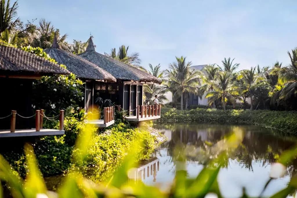 Thatched-roof spa area by a serene pond, surrounded by palms and greenery.